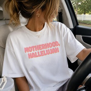 May include: White t-shirt with the text "MOTHERHOOD, HALLELUJAH" in pink and orange lettering. The shirt is being worn in a car. The person is wearing gold hoop earrings and a gold necklace.