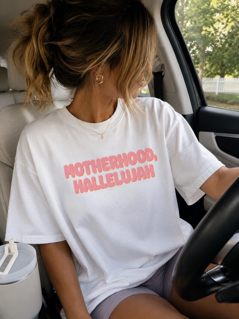 May include: White t-shirt with the text "MOTHERHOOD, HALLELUJAH" in pink and orange lettering. The shirt is being worn in a car. The person is wearing gold hoop earrings and a gold necklace.