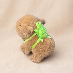 May include: A plush brown capybara toy with a soft, fuzzy texture, featuring a small, bright green turtle-shaped backpack attached with lime green straps. The capybara is positioned at an angle, showing its rounded body, small ears, and closed eyes.