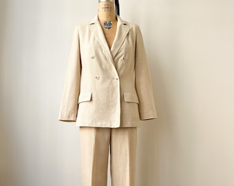 Puede incluir: Un traje de color crema compuesto por una chaqueta de doble botonadura y un pantalón a juego. La chaqueta tiene solapa de muesca y dos botones de cierre. El pantalón tiene pierna recta.