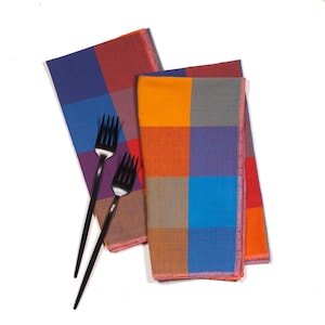 May include: Two checkered napkins in orange, red, blue, and purple. The napkins are folded and stacked on top of each other. Two black forks are placed on top of the napkins.