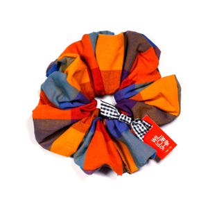 May include: A large, colorful scrunchie with a plaid pattern in shades of orange, blue, red, and brown. It features a small black and white gingham bow and a red tag with white text. The scrunchie is designed for hair styling.