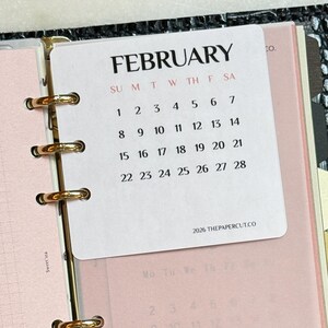 May include: A black textured binder with gold rings holding a white February 2026 calendar card and a translucent pink divider page with the word FEBRUARY printed in large black letters. The calendar displays dates from 1 through 28.