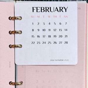 May include: A ring-bound planner featuring a white February 2026 calendar card with black text and red day initials. Beneath it, a translucent pink page displays the word FEBRUARY in large black font. The planner has a black textured cover.