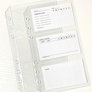 Op de afbeelding: Transparante plastic binder inserts met drie vakken, elk met een witte kaart. De bovenste kaart heeft ruimte voor "Maand" en "Belangrijke datums" met een kalenderraster. De middelste kaart is een "Wekelijkse tracker" met ruimte voor "Stemming" en "Gewoonte".