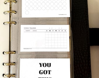 May include: A black ring-bound planner containing three white cards in clear plastic sleeves. The top card features the text I choose myself everyday and a grid of circles. The middle card is a weekly tracker. The bottom card reads YOU GOT THIS.