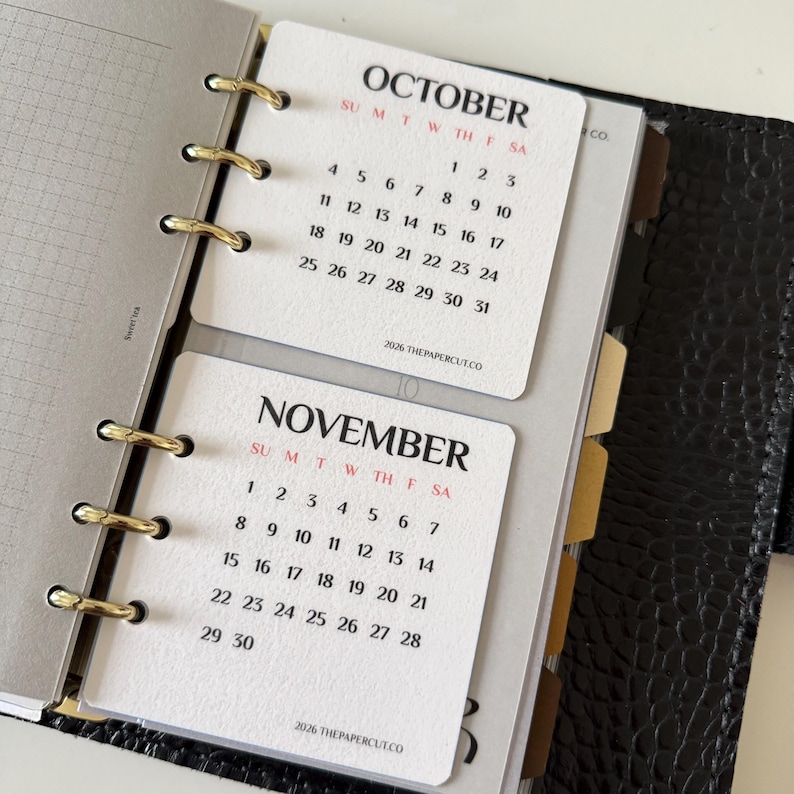 May include: A ring-bound planner featuring white calendar inserts for October and November 2026. The pages display black text with red headers for days of the week, held by gold-coloured metal rings against a black textured cover with tabbed dividers.