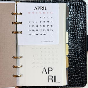 May include: A black textured binder with gold rings holding a white calendar page for April 2026. The page displays the month name, days of the week, and dates 1 through 30, with the text 2026 THEPAPERCUT.CO at the bottom and a secondary April calendar.