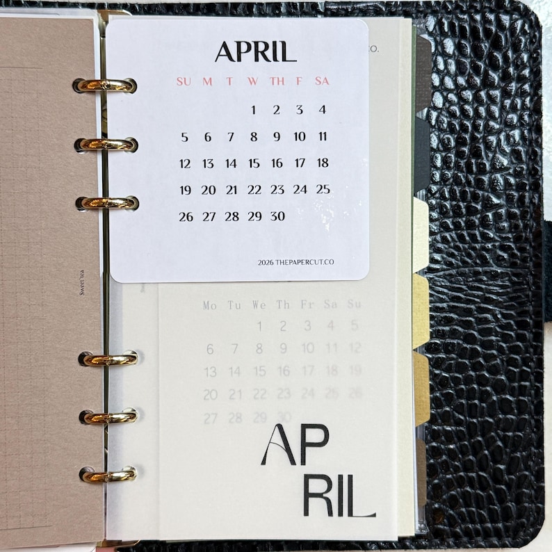 May include: A black textured binder with gold rings holding a white calendar page for April 2026. The page displays the month name, days of the week, and dates 1 through 30, with the text 2026 THEPAPERCUT.CO at the bottom and a secondary April calendar.