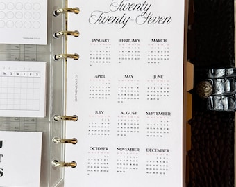 May include: A black leather binder with a six-ring metal mechanism holding a white paper insert. The insert features a 2027 calendar with the text Twenty Twenty-Seven at the top and the months January through December listed in a grid format.