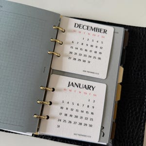 May include: A ring-bound planner featuring cream-coloured calendar inserts for December and January. The pages display black text for the months and dates, with red lettering for the days of the week. Gold-toned metal rings secure the pages.