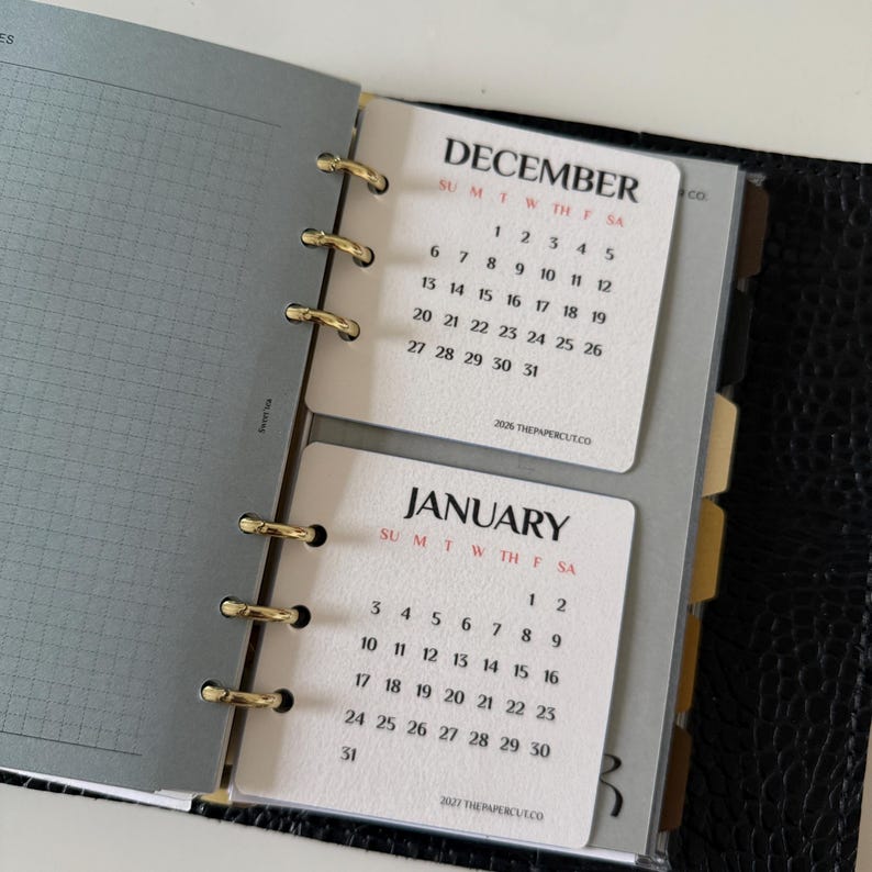 May include: A ring-bound planner featuring cream-coloured calendar inserts for December and January. The pages display black text for the months and dates, with red lettering for the days of the week. Gold-toned metal rings secure the pages.