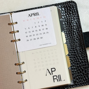 May include: A black textured leather planner with gold rings holding a white April 2026 calendar card. The card displays the month, days of the week, and dates. A translucent page underneath shows a faint calendar grid and the word APRIL in large font.
