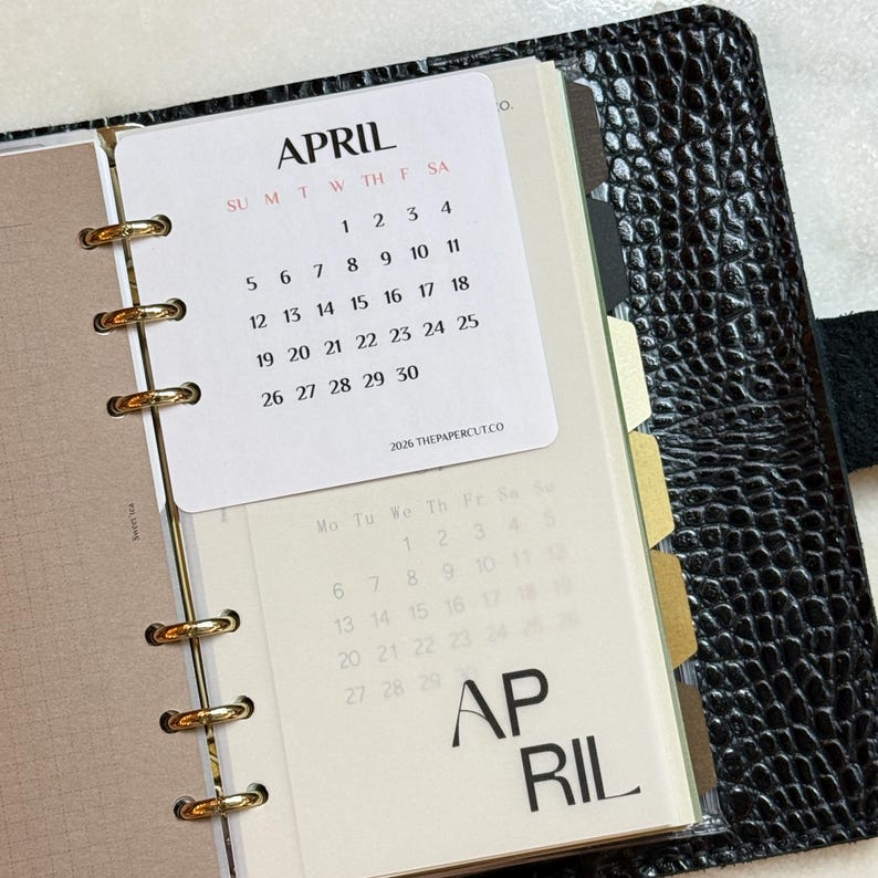 May include: A black textured leather planner with gold rings holding a white April 2026 calendar card. The card displays the month, days of the week, and dates. A translucent page underneath shows a faint calendar grid and the word APRIL in large font.