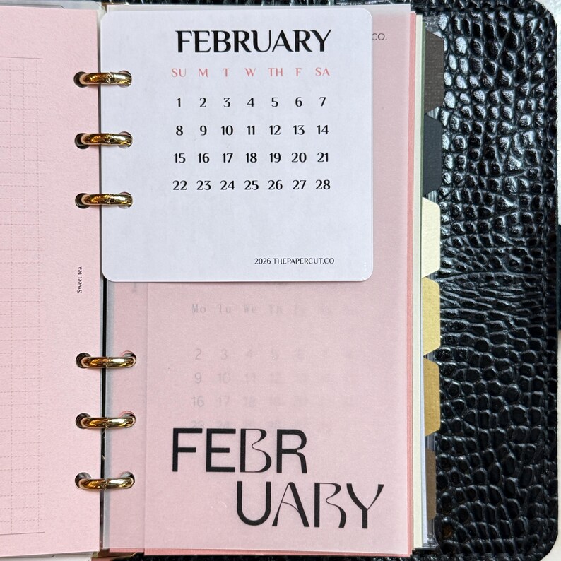 May include: A ring-bound planner featuring a white February 2026 calendar card with black text and red day initials. Beneath it, a translucent pink page displays the word FEBRUARY in large black font. The planner has a black textured cover.