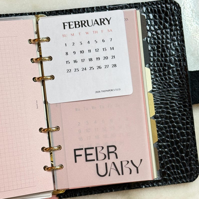 May include: A black textured binder with gold rings holding a white February 2026 calendar card and a translucent pink divider page with the word FEBRUARY printed in large black letters. The calendar displays dates from 1 through 28.