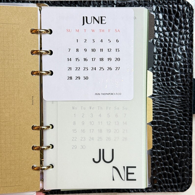 May include: A black textured binder with gold rings holding a June 2026 calendar page. The white page displays the month, days of the week, and dates in black and red text, with the text 2026 THEPAPERCUT.CO printed at the bottom of the calendar.