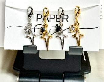 May include: Four star-shaped charms with lobster clasps. Two charms are silver-toned with clear crystals, and two are gold-toned, one with a black gem and one with a clear crystal. The charms are displayed on a white card with the text "PAPER Cut".