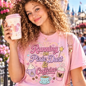 May include: A light pink t-shirt featuring a graphic print with the text Running on Pixie Dust & Caffeine, surrounded by illustrations of coffee cups, hearts, and a star wand. The shirt is paired with blue denim jeans and pink sequined ears.