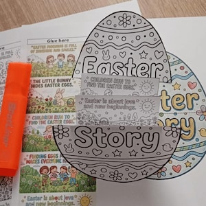 Peut inclure: Un kit de bricolage sur le thème de Pâques avec un livre d'histoires en forme d'œuf. Le kit comprend une page à colorier avec les mots "Easter" et "Story", et des panneaux avec du texte et des illustrations. Un surligneur orange est à gauche.