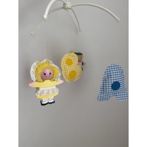 May include: A vintage baby mobile featuring a white frame with hanging fabric figures. The mobile includes a doll with a yellow dress and a bonnet, a yellow gingham "B", and a blue and white gingham "A". The figures are suspended by strings.