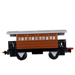 Può includere: Un vagone di treno giocattolo marrone con tetto grigio e ruote nere. Il vagone ha piccole finestre con immagini di persone all'interno. Il vagone del treno giocattolo è un design classico.