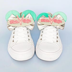 May include: A pair of white lace-up sneakers featuring decorative attachments on the tongues. Each attachment includes two mint green circular ears, a metallic silver bow, and an oval patch with the pink embroidered text Flo's Cafe.