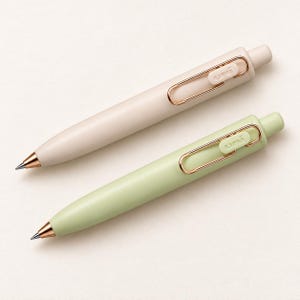 Könnte beinhalten: Zwei einziehbare Kugelschreiber mit mattem Gehäuse in Beige und Hellgrün. Jeder Stift verfügt über einen roségoldfarbenen Metallclip in Büroklammerform, eine passende roségoldfarbene Spitze und das Wort uni auf dem Clip.