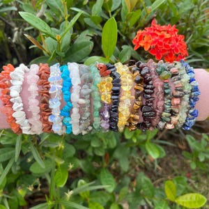 May include: A collection of gemstone chip bracelets in various colors, including orange, white, pink, brown, blue, green, yellow, black, and purple. The bracelets are displayed on a pink cylindrical stand, surrounded by green foliage and red flowers.