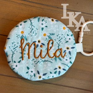 May include: A round, light blue baby toy with a floral pattern featuring white and yellow flowers and green leaves. The name "Mila" is embroidered in a gold-colored thread. A white plastic ring and ribbon are attached.