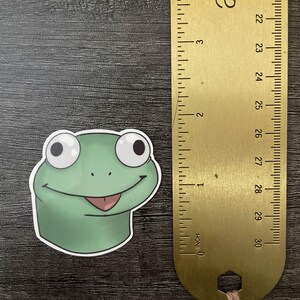 Może przedstawiać: Zielona naklejka z żabą z uśmiechniętą twarzą i r&oacute;żowym językiem. Złota linijka obok naklejki mierzy do 7,6 cm. Naklejka znajduje się na ciemnej drewnianej powierzchni.