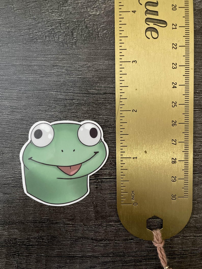 Może przedstawiać: Zielona naklejka z żabą z uśmiechniętą twarzą i r&oacute;żowym językiem. Złota linijka obok naklejki mierzy do 7,6 cm. Naklejka znajduje się na ciemnej drewnianej powierzchni.