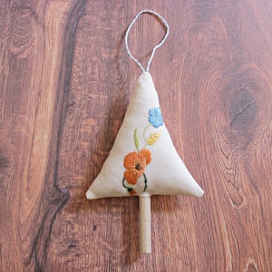 Könnte beinhalten: Ein weißer Stoff-Weihnachtsbaumschmuck mit gestickten orangefarbenen und blauen Blumen. Der Schmuck hat einen Holzstab zum Aufhängen.