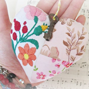 May include: A handmade heart-shaped pillow with a patchwork design. The pillow is made of pink, white, and brown fabric and features embroidered flowers and leaves. A small antique key hangs from the pillow.