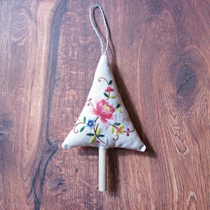 May include: A white fabric Christmas tree ornament with embroidered flowers in pink, red, yellow, blue, and green. The tree has a wooden dowel for a trunk.