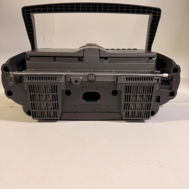 K&ouml;nnte beinhalten: Ein dunkelgrauer, tragbarer Ghettoblaster mit einem oben angebrachten Tragegriff, einer horizontalen Teleskopantenne und zwei Lautsprechergittern auf der R&uuml;ckseite. Das Ger&auml;t verf&uuml;gt &uuml;ber ein Kunststoffgeh&auml;use mit sichtbaren L&uuml;ftungsschlitzen.