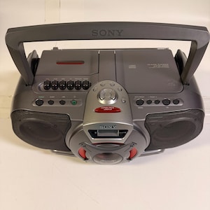 K&ouml;nnte beinhalten: Ein dunkelgrauer tragbarer Sony-Boombox mit CD-Player, Kassettendeck und zentralem Power Drive Woofer. Das Ger&auml;t verf&uuml;gt &uuml;ber einen gro&szlig;en Tragegriff, zwei seitliche Lautsprecher mit schwarzen Gittern und verschiedene Bedientasten.