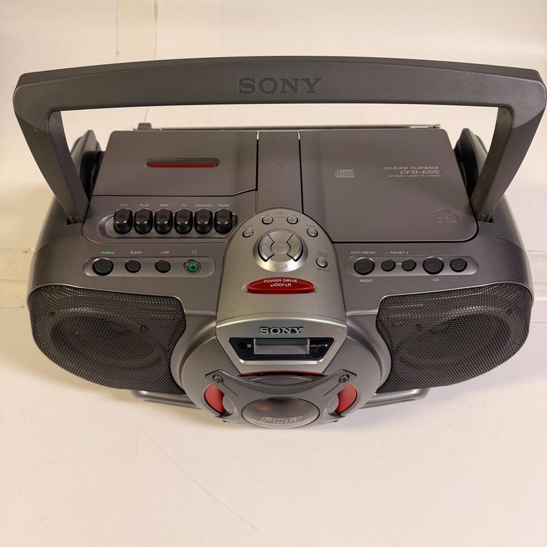 K&ouml;nnte beinhalten: Ein dunkelgrauer tragbarer Sony-Boombox mit CD-Player, Kassettendeck und zentralem Power Drive Woofer. Das Ger&auml;t verf&uuml;gt &uuml;ber einen gro&szlig;en Tragegriff, zwei seitliche Lautsprecher mit schwarzen Gittern und verschiedene Bedientasten.