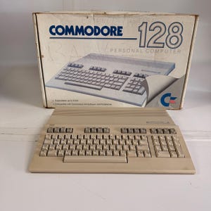 May include: A vintage Commodore 128 personal computer with its original box. The beige keyboard is in the foreground, with the box behind it. The box features the Commodore logo and the text "128 PERSONAL COMPUTER".
