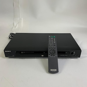Peut inclure: Un lecteur DVD Sony noir au châssis rectangulaire, doté d'un tiroir à disque central et d'un bouton d'alimentation sur la gauche. Une télécommande noire assortie avec de nombreux boutons, dont un pavé de navigation, est posée dessus.