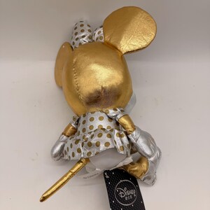 May include: A plush Minnie Mouse doll in a gold and silver color scheme. The doll features a gold head, ears, and bow with silver polka dots. The dress is silver with gold polka dots. The doll has a gold tail and silver shoes.