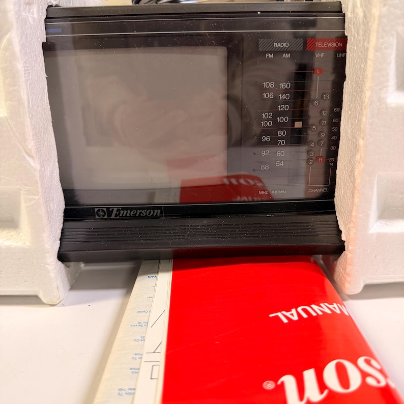 K&ouml;nnte beinhalten: Ein schwarzes, tragbares Emerson-Fernseher- und Radioger&auml;t aus den 1980er Jahren. Das Ger&auml;t hat einen kleinen Bildschirm, einen Kanalwahlschalter und Radiofrequenzanzeigen. Ein rotes Handbuch ist unter dem Ger&auml;t sichtbar, verpackt in wei&szlig;em Schaumstoff.