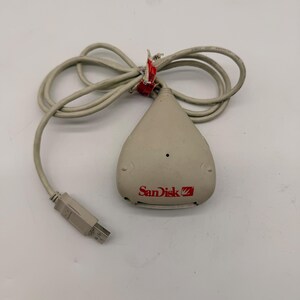 May include: A beige Sandisk card reader with a USB connector and a grey cable. The Sandisk logo is printed in red on the device. The cable is secured with red tape.