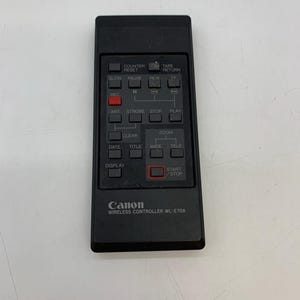 Könnte beinhalten: Eine schwarze Canon Wireless Controller WL-E708. Die Fernbedienung hat mehrere Tasten mit Aufschriften wie "REC", "PAUSE", "ZOOM" und "START/STOP". Das Canon-Logo ist unten auf der Fernbedienung zu sehen.