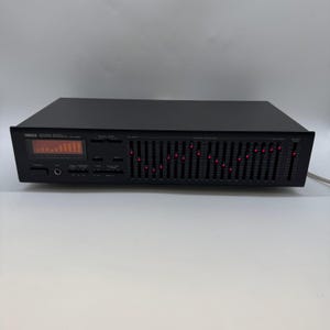 Può includere: Equalizzatore grafico Yamaha EQ-500 nero con display digitale e numerosi cursori. L'unità ha un design elegante con spie rosse e un cavo di alimentazione.