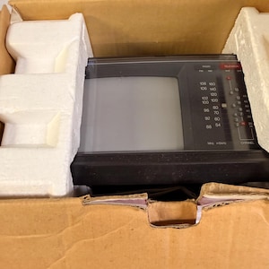 K&ouml;nnte beinhalten: Ein alter, tragbarer Fernseher in Schwarz und Grau, eingebettet in seinem originalen Karton mit sch&uuml;tzenden Schaumstoffeins&auml;tzen. Der Fernseher hat einen kleinen Bildschirm, einen Kanalwahlschalter und Radiofrequenzanzeigen. Die Box ist braun.