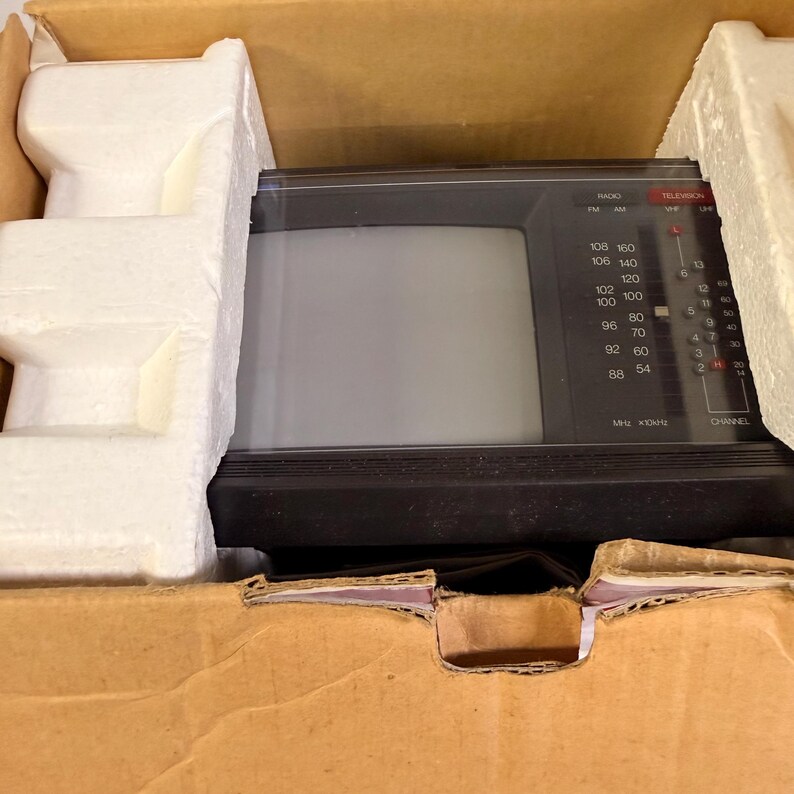 K&ouml;nnte beinhalten: Ein alter, tragbarer Fernseher in Schwarz und Grau, eingebettet in seinem originalen Karton mit sch&uuml;tzenden Schaumstoffeins&auml;tzen. Der Fernseher hat einen kleinen Bildschirm, einen Kanalwahlschalter und Radiofrequenzanzeigen. Die Box ist braun.