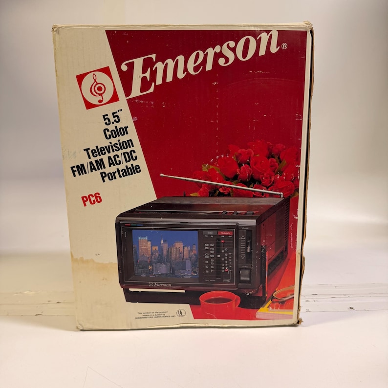 K&ouml;nnte beinhalten: Ein Vintage Emerson 5,5" Farbfernseher, FM/AM AC/DC tragbar, Modell PC6, in der Originalverpackung. Die Box hat ein rot-wei&szlig;es Design mit dem Emerson-Logo und einer Grafik des Fernsehers. Der Fernseher zeigt eine Stadtlandschaft.