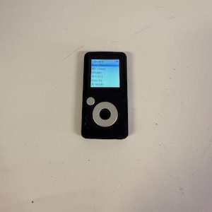 May include: A black digital music player with a rectangular screen displaying a menu with the text Library, Now playing, All songs, Albums, Artists, Genres, and Browser. The device features a circular silver navigation wheel and a small button.