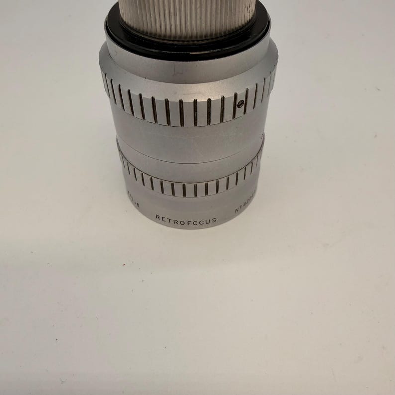 May include: A silver-colored camera lens with the word "RETROFOCUS" printed on the side. The lens has a ridged design and a black top. The lens is cylindrical and sits on a white surface.